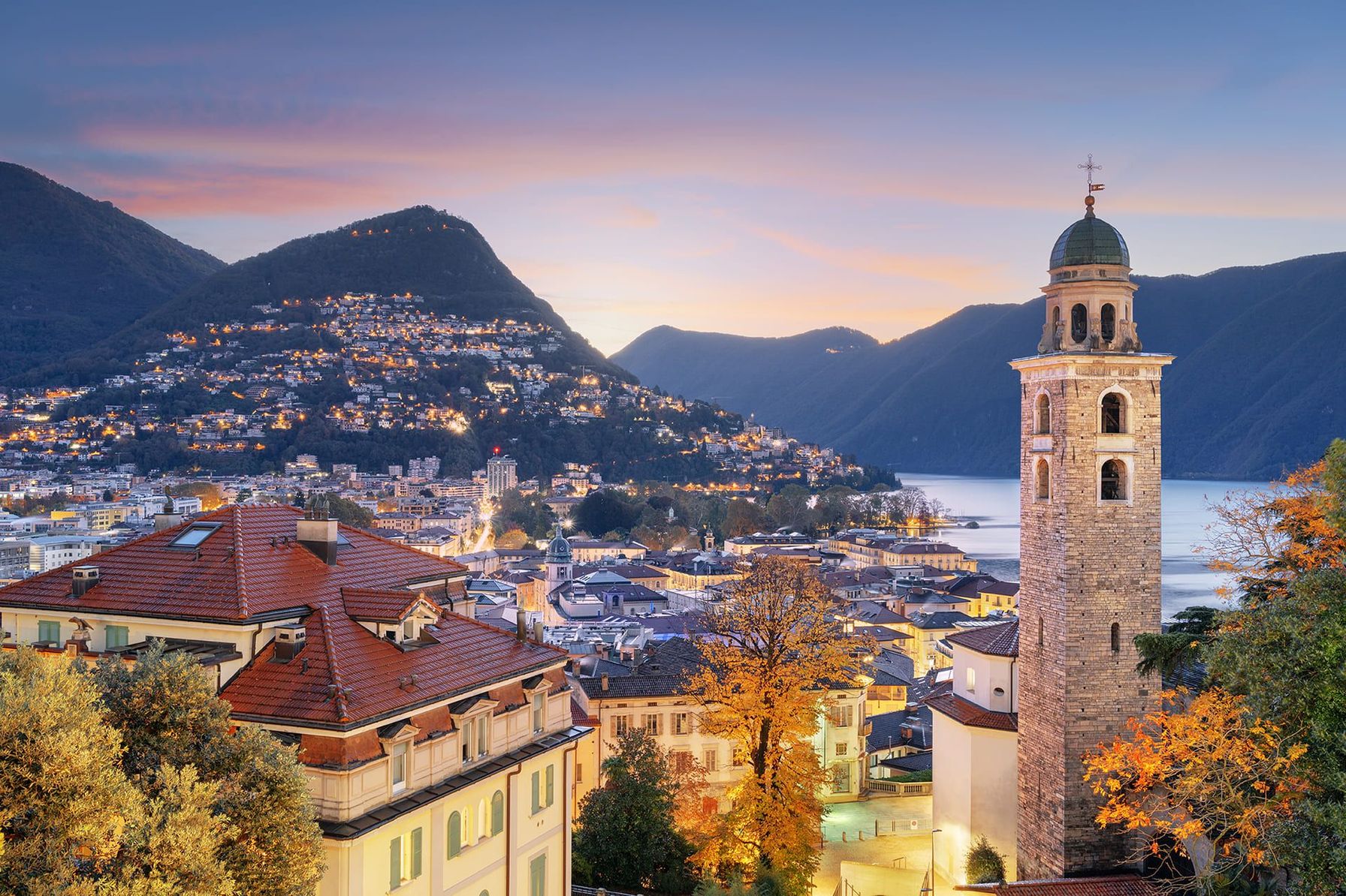 The breathtaking beauty of Lugano at sunset, with its illuminated hillside, historic architecture, and serene lake views.