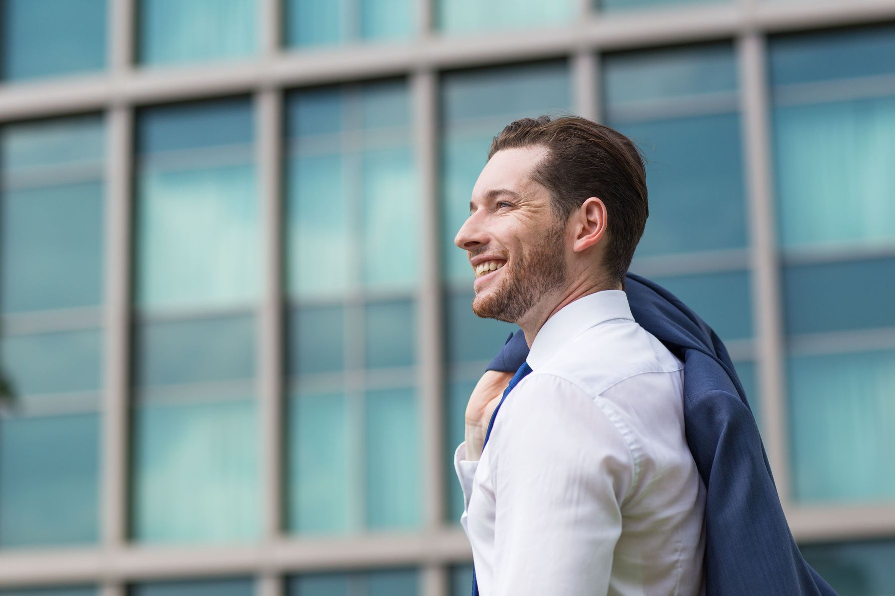 A cheerful businessman in a white shirt carrying his jacket over his shoulder, standing outside a modern glass office building.