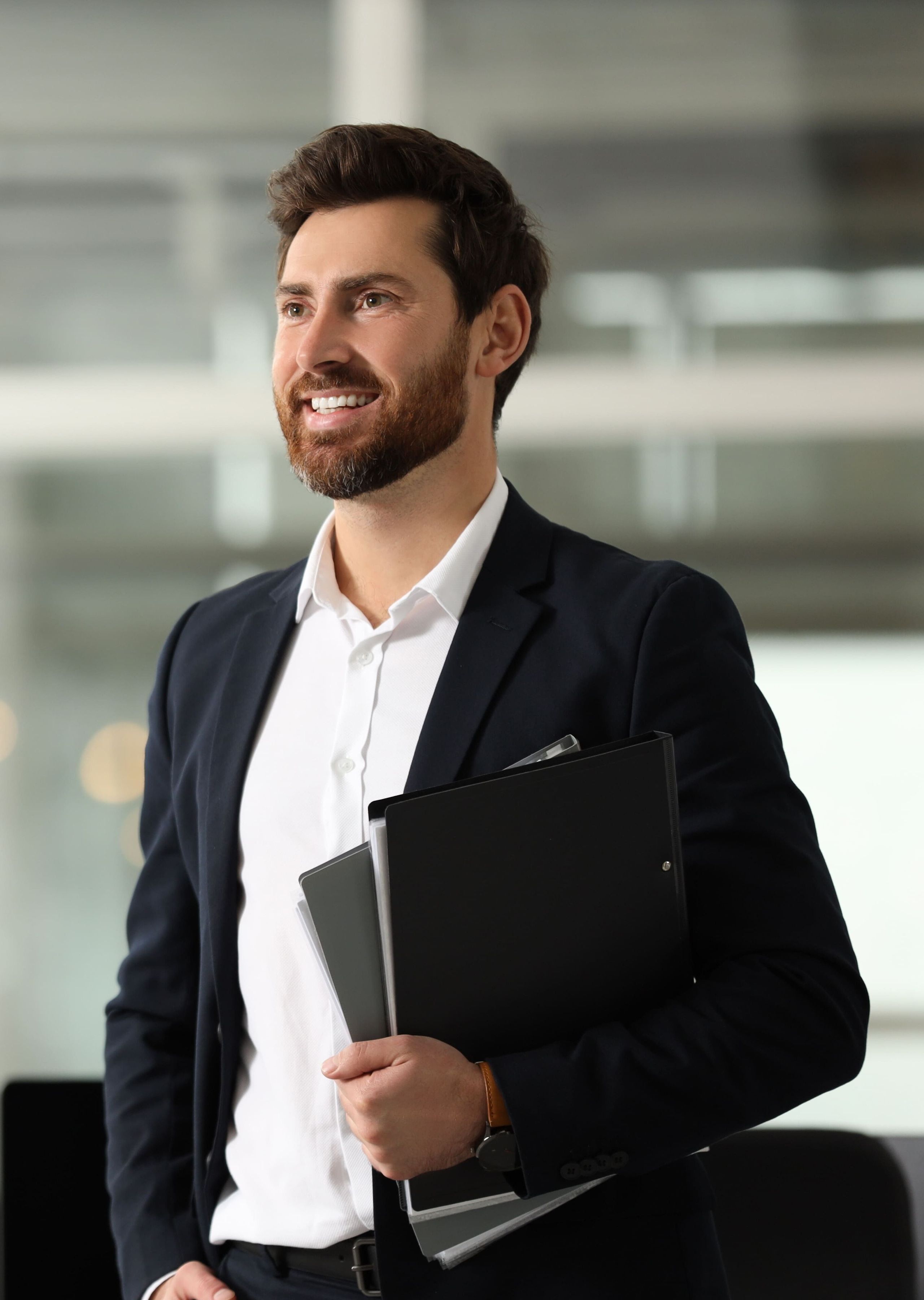 A smiling businessman in a formal suit holding documents and a tablet, standing in a bright, modern office.